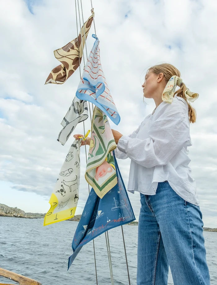 En tjej står bredvid några färgglada sjalar som fladdrar i vinden, uppknutna på ett stag på en båt. Havet syns i bakgrunden.