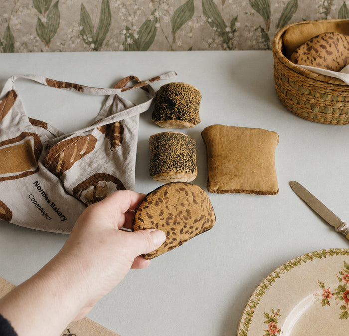 Person holding a loaf of play food bread with a basket of bread and a floral plate on a table.