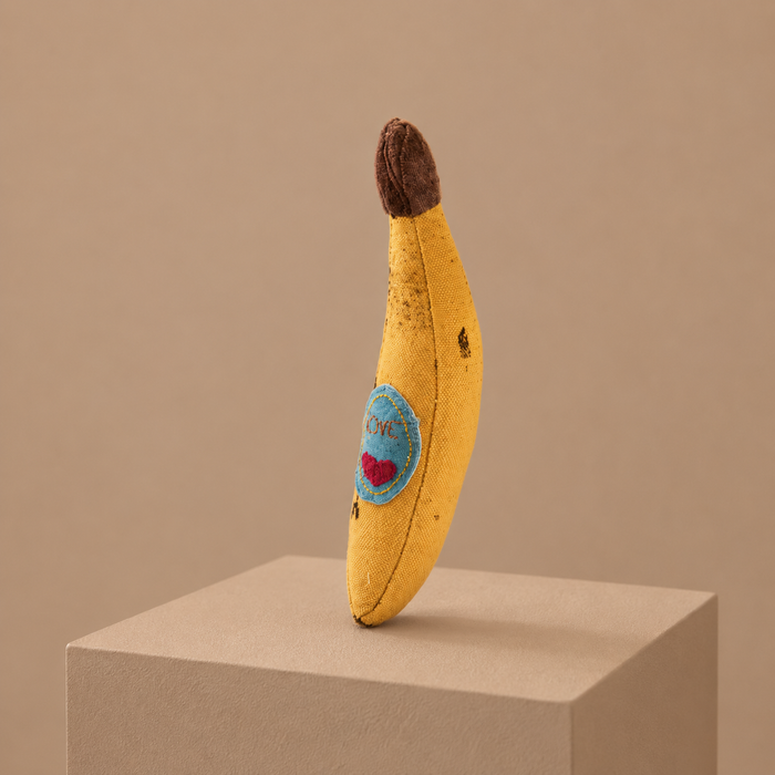 Handmade fabric banana toy with embroidered love patch, displayed upright on a neutral studio pedestal.