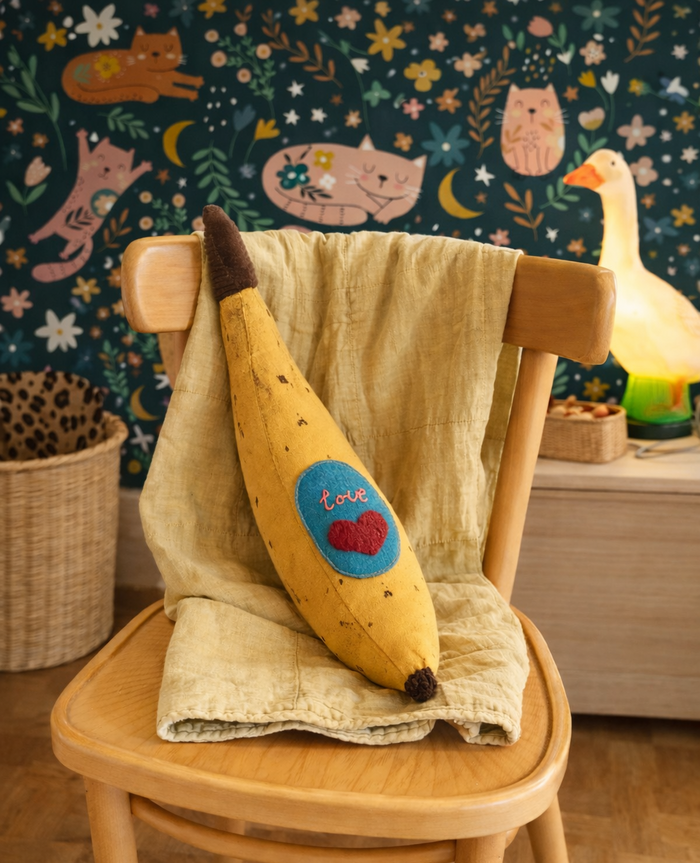 Banana-shaped canvas pillow with a heart and 'love' text on a chair against a colorful wall.