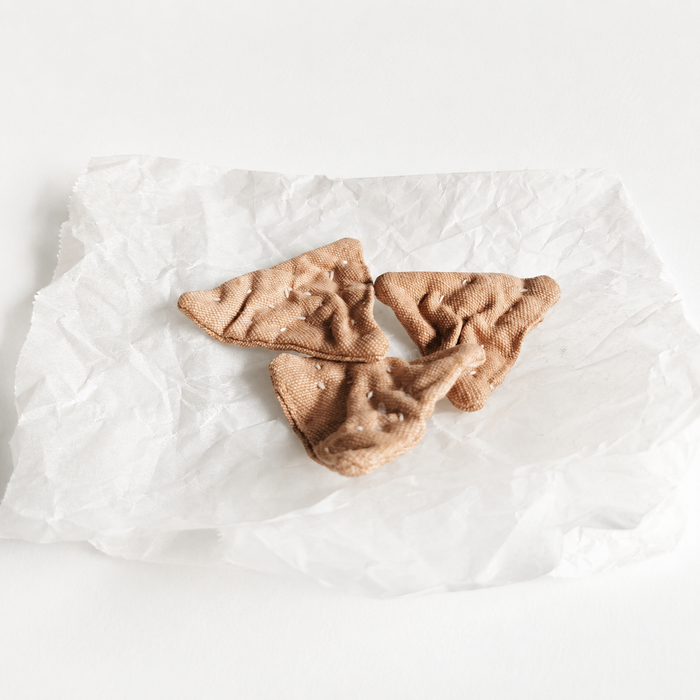 Handmade fabric triangle crackers displayed on crumpled white paper in a light studio product setting. Soft textile pretend play food with hand-printed details and small embroidered seeds.