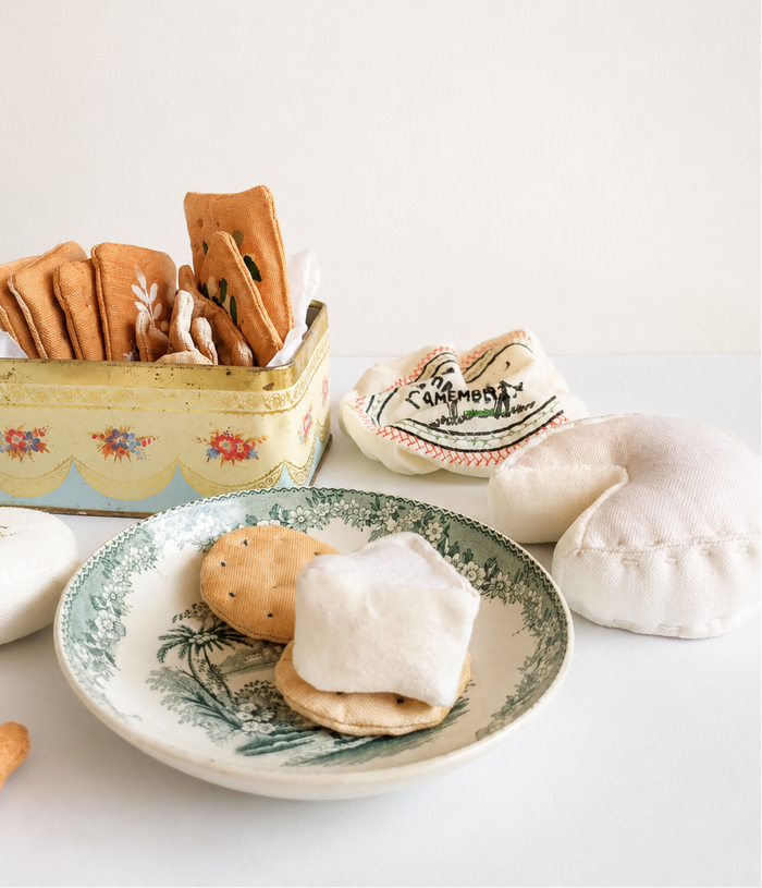 Handmade fabric play food set with whole wheat crackers, triangle crackers and soft textile cheese displayed in a vintage tin and on a floral plate on a white tabletop against a plain wall.