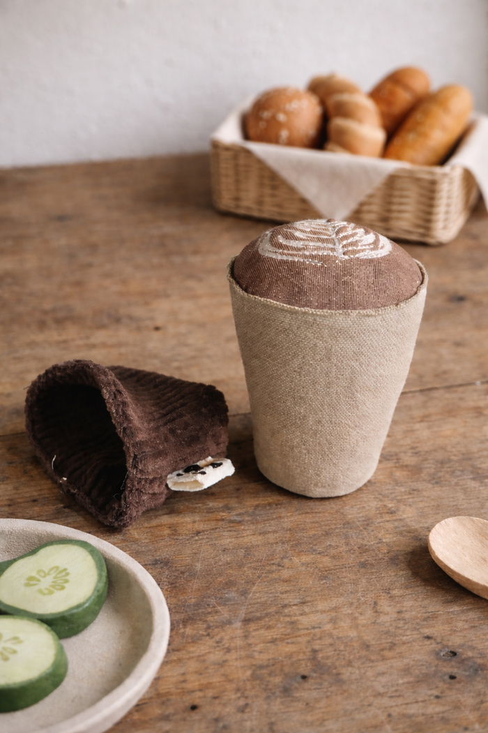 Handmade fabric cappuccino cup play food styled on a rustic wooden table with pretend food toys and a bread basket in the background.