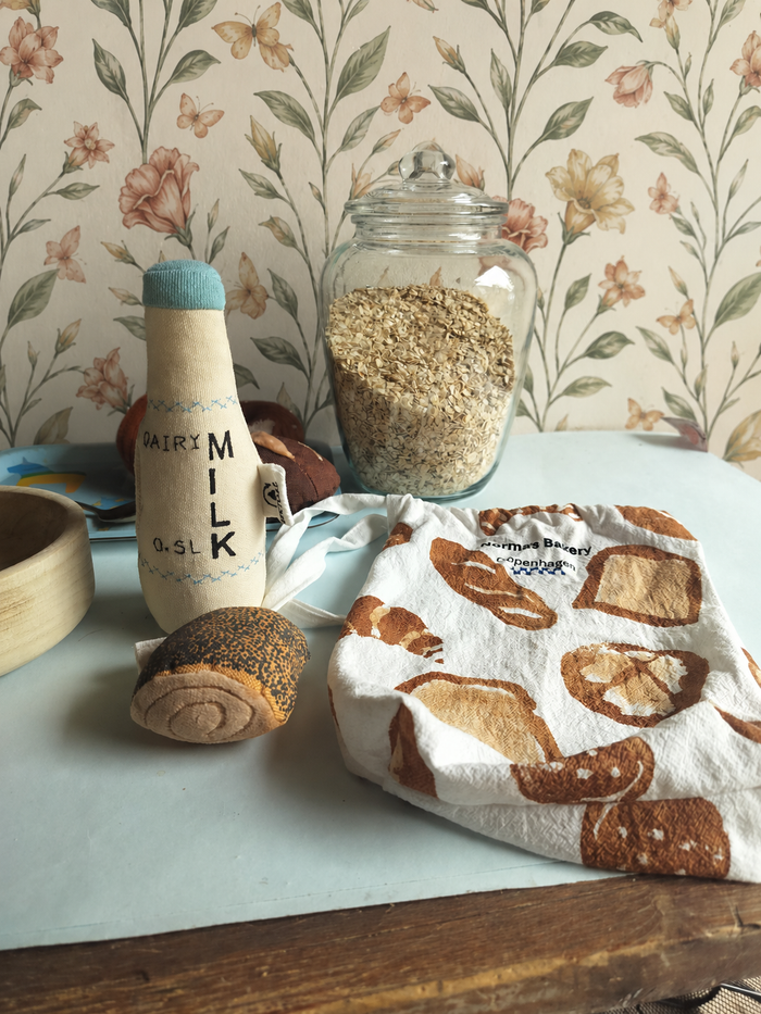 Play Food Milk bottle, jar of oats, and bread-themed towel on a table with floral wallpaper.