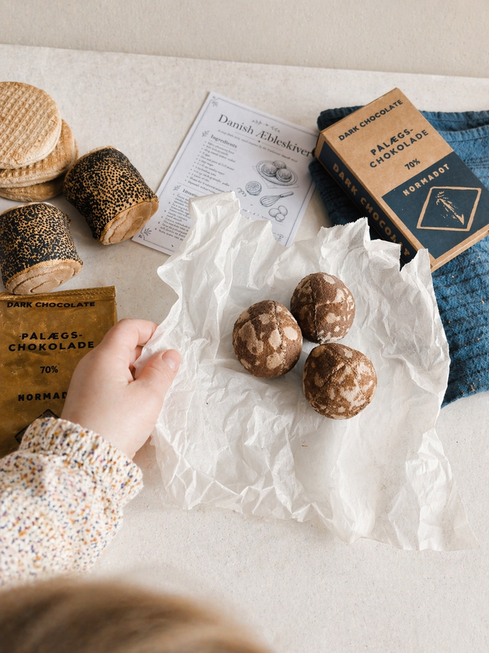 Child playing with handmade bakery play food, including fabric æbleskiver, stroopwafels, chocolate and a recipe card on a light table