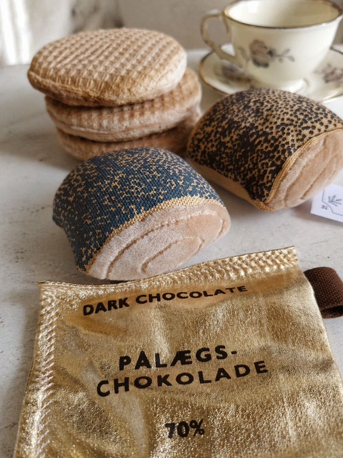 Close-up of handmade bakery play food with fabric stroopwafels, bread rolls and dark chocolate in a soft café-inspired setting.