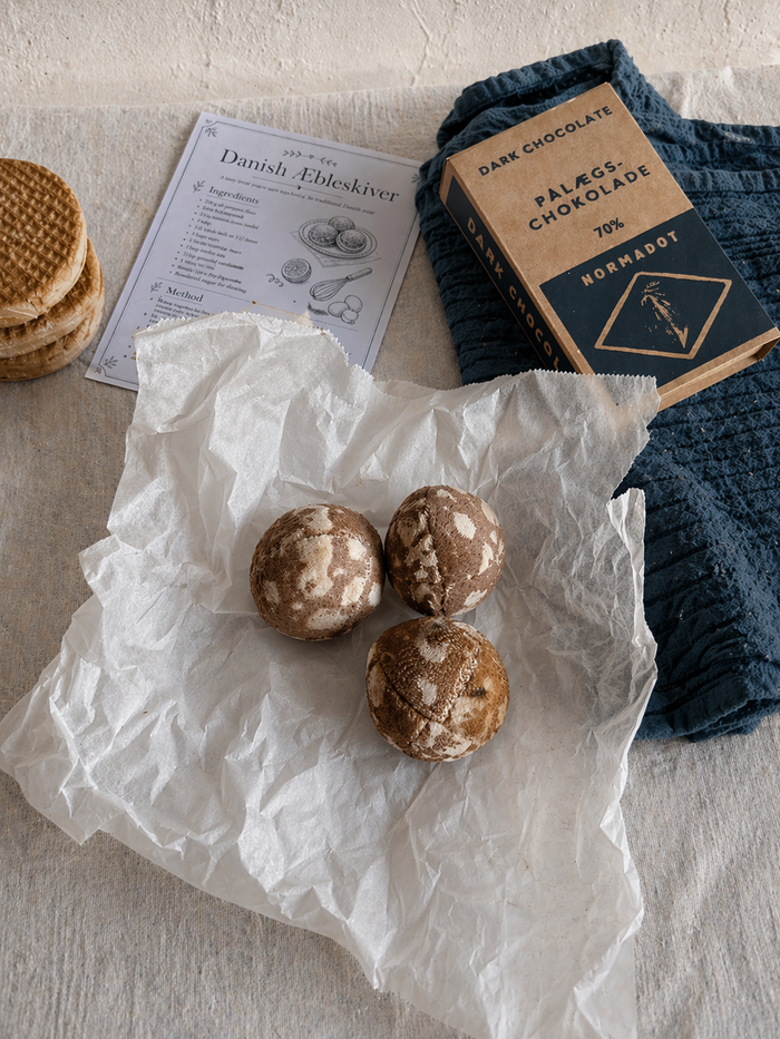 Handmade bakery play food with fabric æbleskiver on tissue paper, stroopwafels, chocolate and a recipe card in a soft tabletop setting.