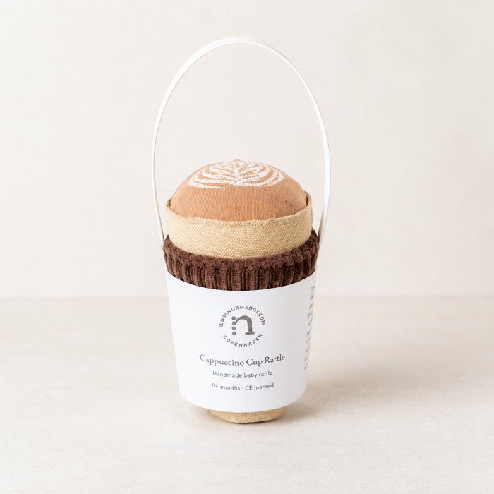 Cappuccino cup rattle with brown velvet band in a white packaging on a light beige background