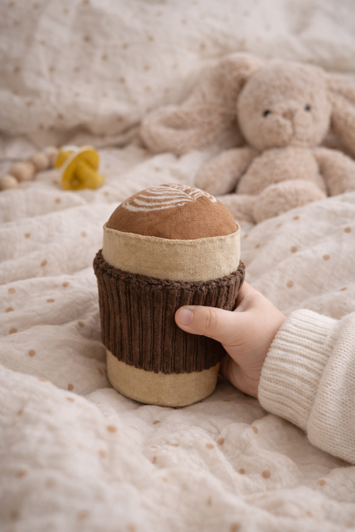 Baby holding handmade cappuccino cup rattle on soft blanket with teddy bear in background