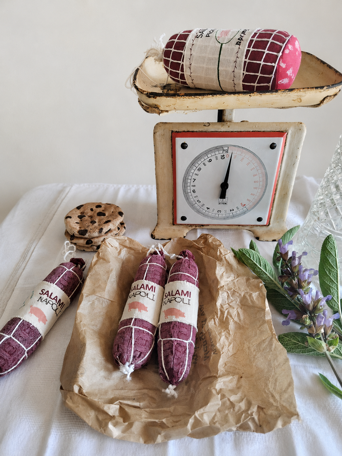 Handmade fabric Salami Napoli play food with printed textile labels, white thread details and recycled polyester filling, styled on brown deli paper with a vintage kitchen scale, cookies and fresh herbs.
