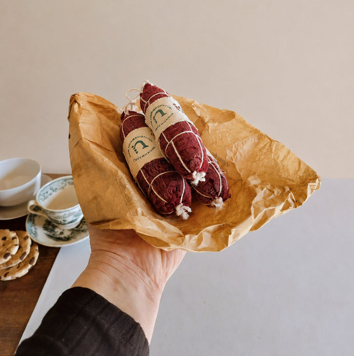 Hand holding handmade fabric Salami Napoli play food wrapped in brown deli paper, styled with vintage cups and textile cookies in a soft neutral product setting.