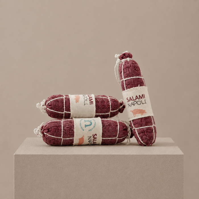 Handmade fabric Salami Napoli play food with printed textile labels, white thread details and recycled polyester filling, displayed on a neutral museum-style pedestal in a soft studio setting