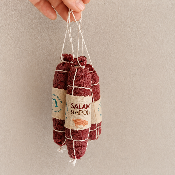 Handmade fabric Salami Napoli play food hanging by white string against a neutral wall.