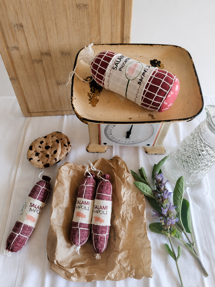 Handmade fabric Salami Napoli play food styled with a vintage kitchen scale, wooden cutting board, textile cookies, brown deli paper and fresh herbs in a rustic product setting