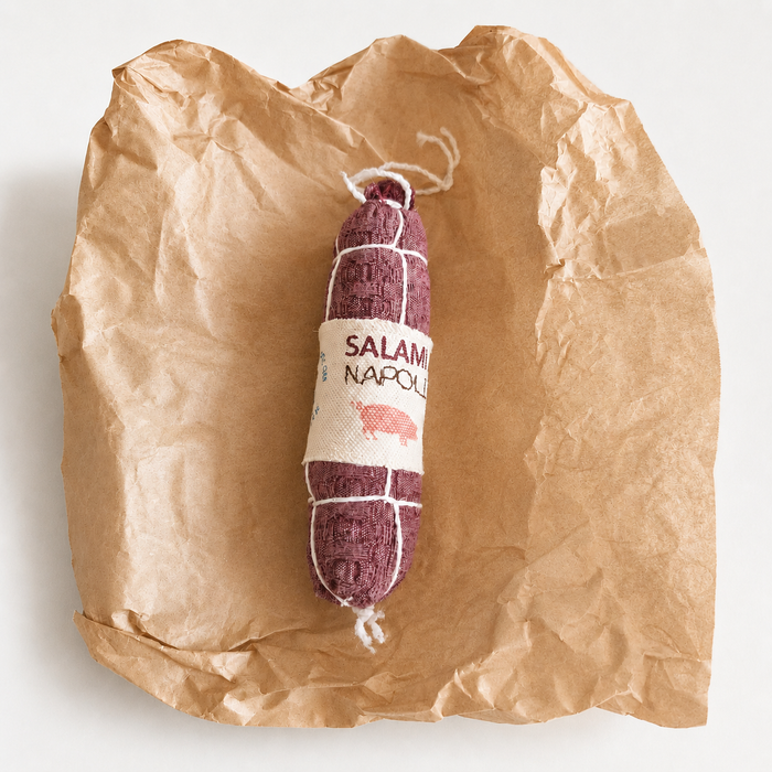 Handmade fabric Salami Napoli play food made from waffle woven cotton with printed textile label, white thread details and recycled polyester filling, displayed on crinkled brown deli paper.