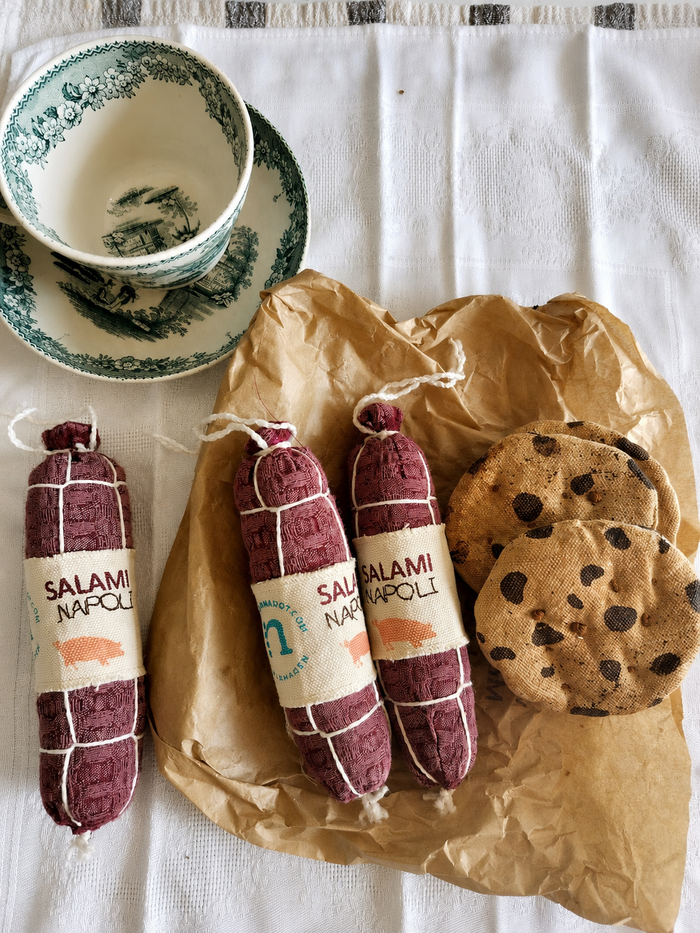 Handmade fabric Salami Napoli play food styled with textile cookies, a vintage cup and brown deli paper on a white cloth in a rustic product setting.