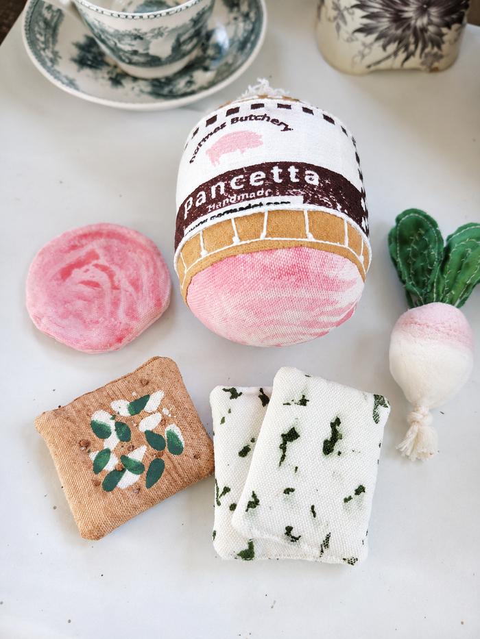 Handmade fabric Pancetta play food with pancetta slice, radish, crackers and cheese pieces in a natural tabletop product setting.
