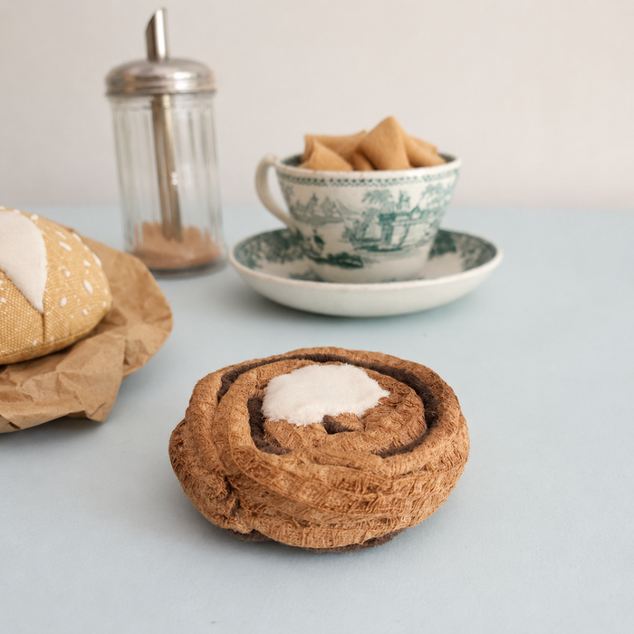 Handmade Danish kanelsnegl fabric play food with velour icing, styled with a tea cup, cinnamon shaker and fabric bread roll.
