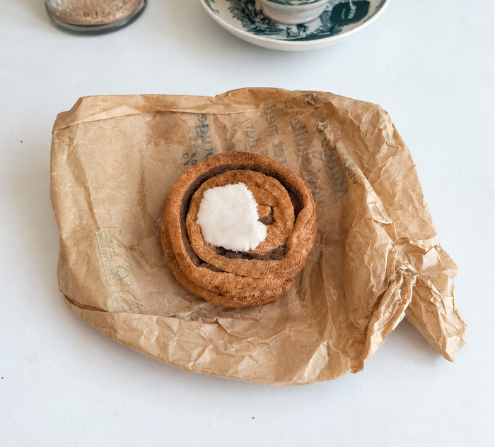 Overhead view of a handmade Danish kanelsnegl fabric play food with velour icing, displayed on crumpled brown paper.
