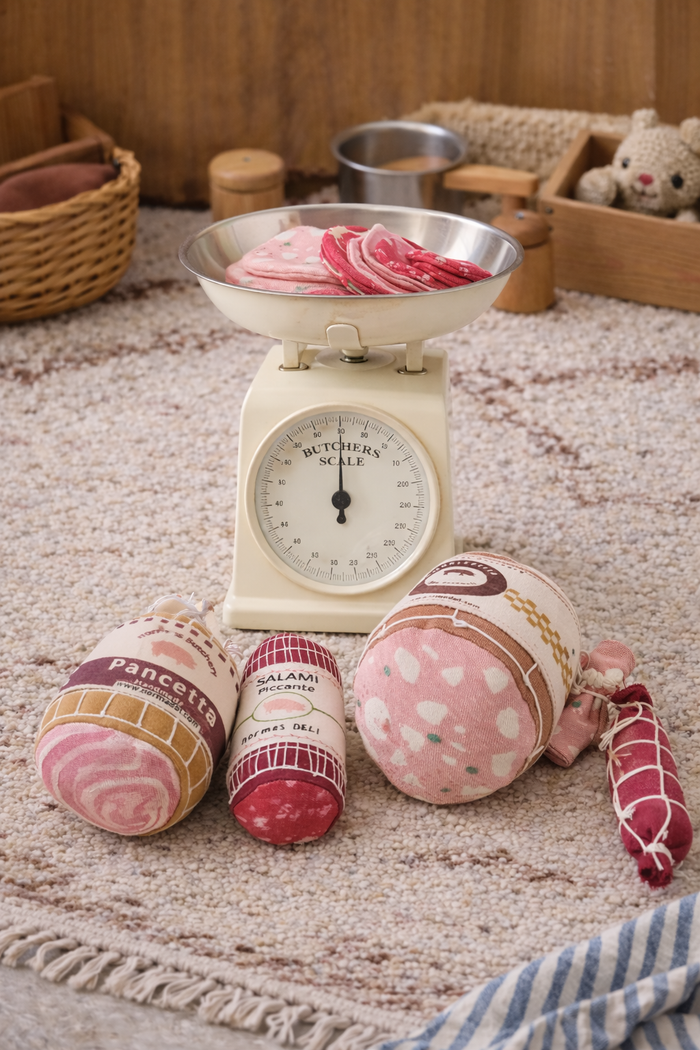 Handmade fabric charcuterie play food set with salami, pancetta and mortadella toys arranged beside a vintage scale in a cozy playroom setting.