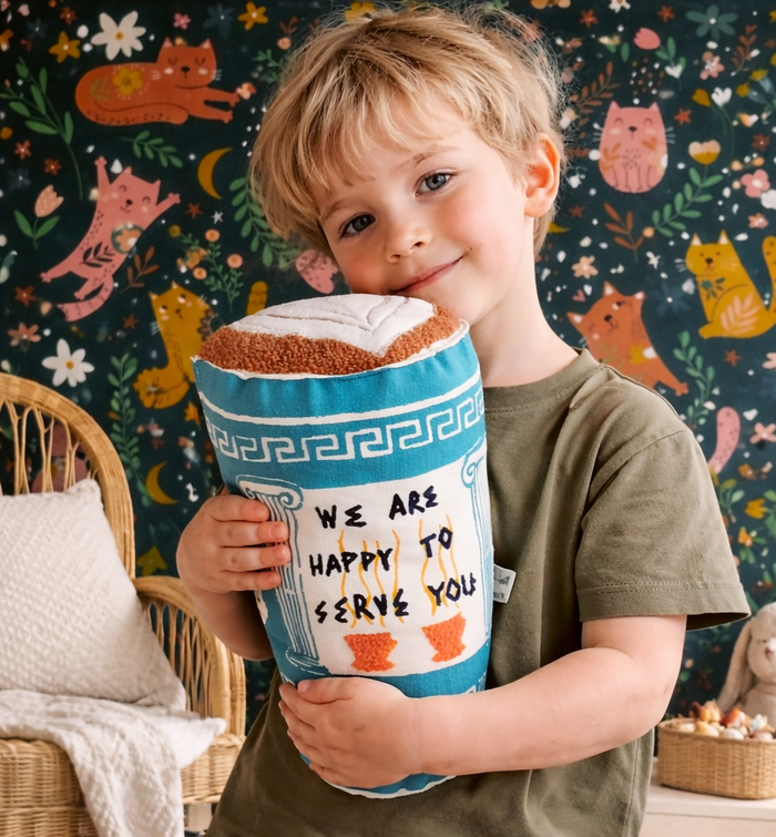 Child holding a toy coffee cup pillow with a colorful cat-themed wallpaper in the background
