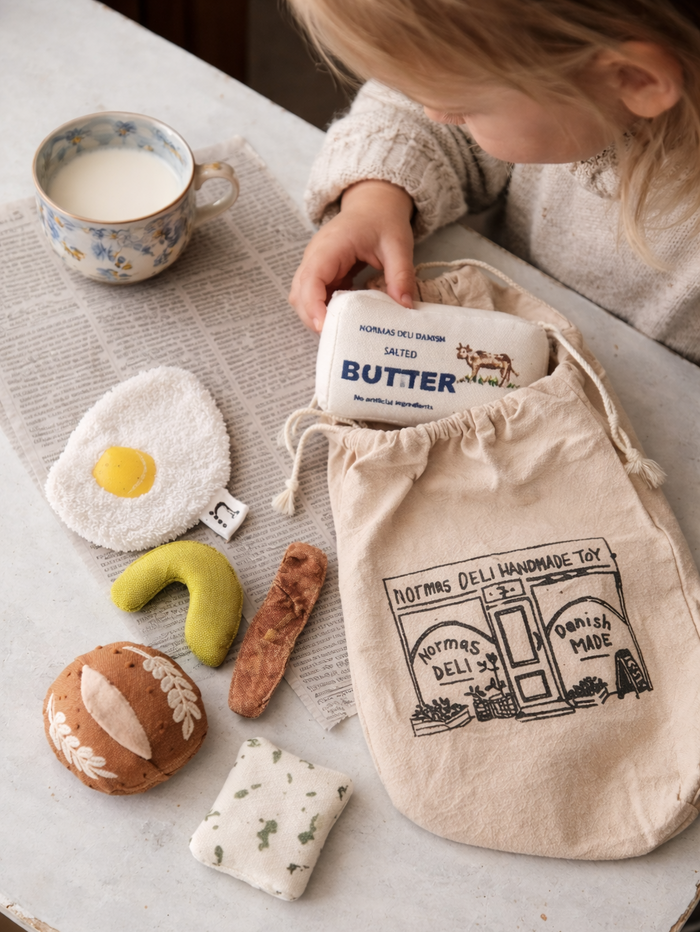 Handmade morning breakfast play food set with soft fabric fried egg, bacon, butter, cucumber, bread roll and cotton deli bag styled on a table.