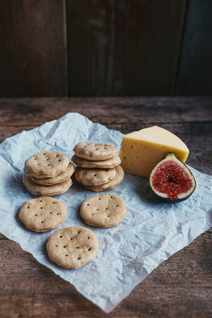 Stacks of fabric play food crackers with a wedge of cheese and fig on a wooden surface